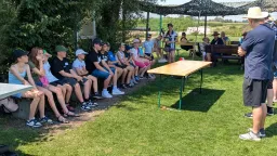 Mehrere Kinder und Jugendliche sitzen auf Bänken unter einem Pavillon, ein Erwachsener steht rechts und hält einen Vortrag