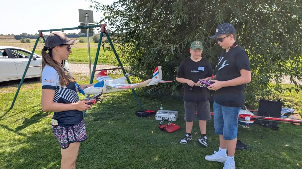 Zwei Jugendliche und ein Kind stehen auf einer Wiese mit Modellflugzeugen, Fernsteuerungen und Ausrüstung