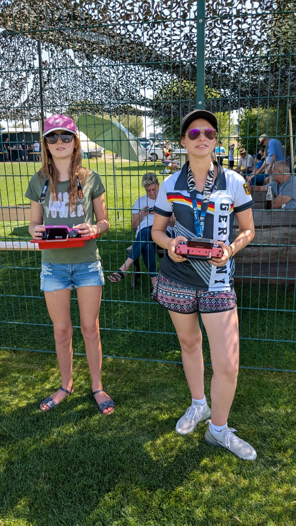 Zwei junge Frauen in Sportkleidung stehen auf Rasen, halten Modellflug-Fernsteuerungen, Zaun im Hintergrund