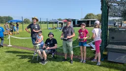 Mehrere Personen verschiedenen Alters an einem Modellflugplatz mit Kunstrasenfeld, Vereinsgelände und Pavillons im Hintergrund