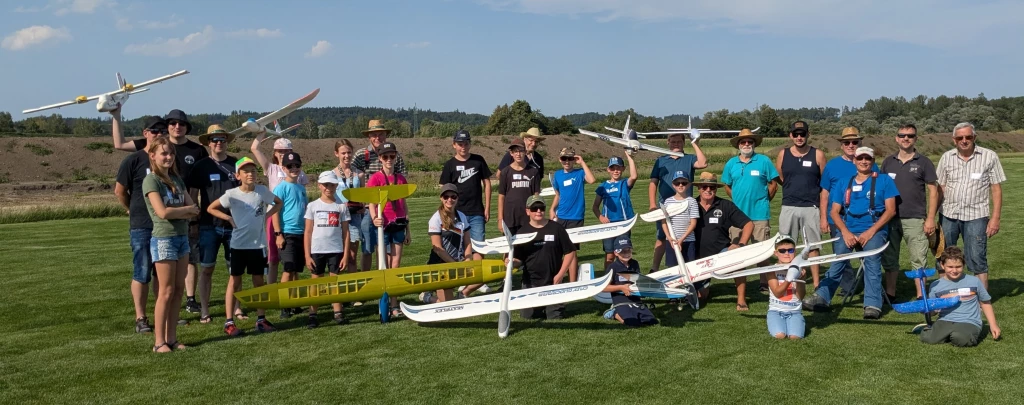 Große Gruppe von Personen unterschiedlichen Alters mit Modellflugzeugen auf Grünfläche, verschiedene weiße und gelbe Flugmodelle
