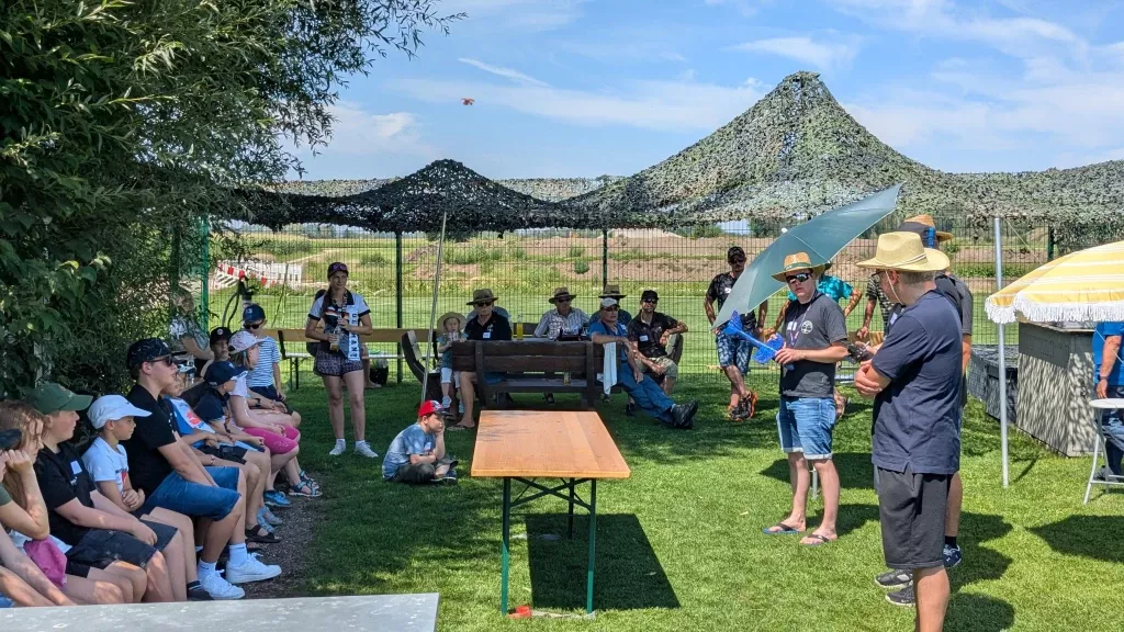 Gruppe von Menschen unter Sonnenschirmen auf Rasenfläche, Berglandschaft im Hintergrund, Modellflug-Veranstaltung