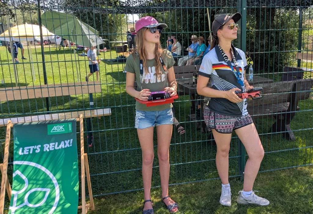 Zwei junge Frauen mit Fernsteuerungen an einem Modellflug-Event, AOK-Banner 'Let's relax', Zeltgelände im Hintergrund