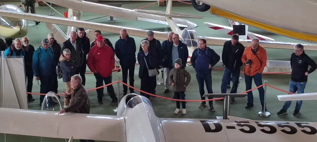 Große Gruppe von Personen verschiedenen Alters in einem Flugzeugmuseum, umgeben von Segelflugzeugen und einer roten Absperrung
