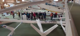 Mehrere weiße Segelflugzeugmodelle in einem Ausstellungsraum mit Besuchergruppe und Absperrungen