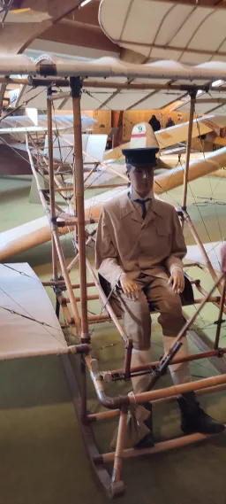 Museale Ausstellung eines historischen Flugzeugs mit Mannequin in Piloten-Uniform, Holzgestell und Flügel im Hintergrund