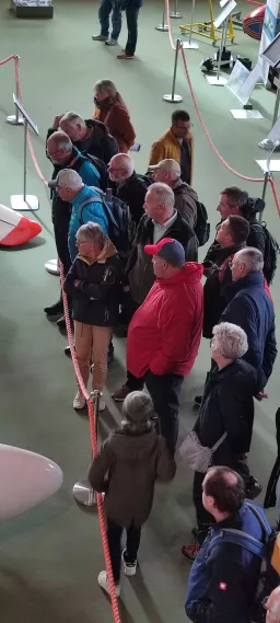 Mehrere ältere Personen stehen in einer Reihe hinter einem roten Absperrband in einem grünen Ausstellungsraum