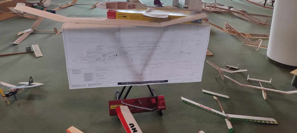 Großformatige technische Bauzeichnung eines Flugmodells auf einem Stativ, umgeben von verschiedenen angefangenen Modellbau-Holzteilen auf grünem Untergrund