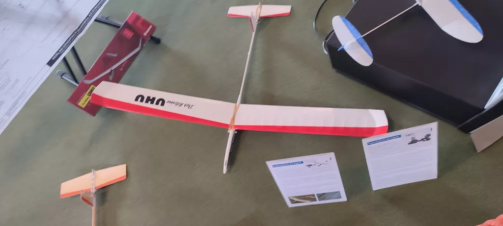 Holz-Modellflugzeug in rot-weiß mit Bauanleitungsblättern auf grünem Untergrund, mehrere Flugzeugteile