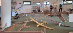 Mehrere Holz-Modellflugzeuge in verschiedenen Größen und Farben auf grünem Boden, im Hintergrund Informationstafeln und ein Mensch