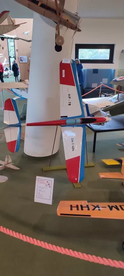 Detailaufnahme eines rot-weiß-blauen Modellflugzeugs, ausgestellt in einem Raum mit Fenster und weiteren Flugzeugmodellen im Hintergrund