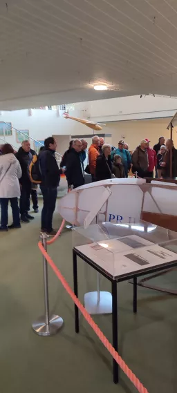 Gruppe älterer Herren in Ausstellungsraum, im Vordergrund Modellflugzeug-Vitrine mit Absperrband, Ausstellungsraum mit weißer Decke