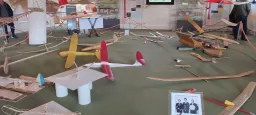 Zahlreiche Modellflugzeuge auf grünem Boden, verschiedene Holz- und Kunststoffmodelle, rote Absperrung, Ausstellungsraum mit Vitrinen