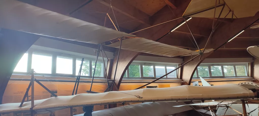 Blick in einen Museumsraum mit historischen Segelflugzeugen, Holzbalken und Panoramafenstern mit Baumblick
