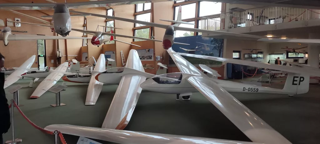 Mehrere weiße Segelflugzeugmodelle in einem Innenraum mit Ausstellungscharakter, grüner Bodenfläche und Absperrband