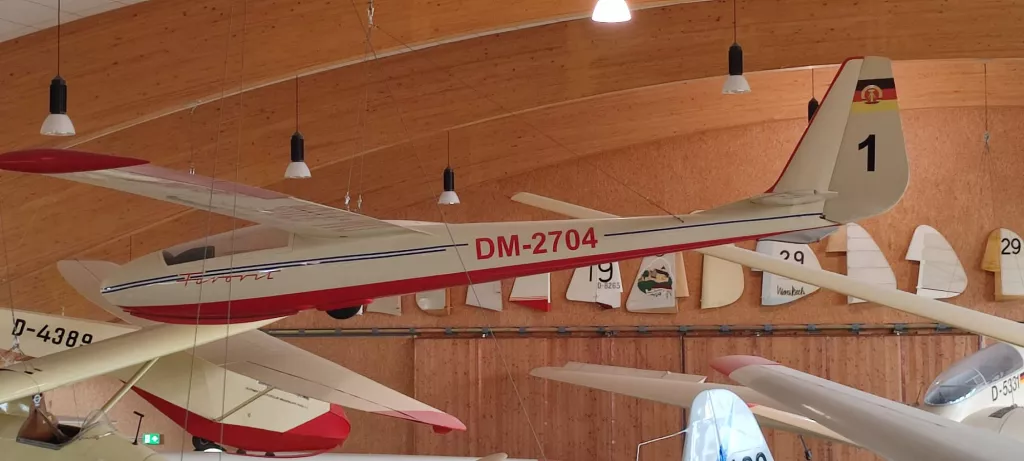 Weißes und rotes Segelflugmodell vom Typ DM-2704 mit Kennzeichen, in Holzhalle ausgestellt, hängt an Decke