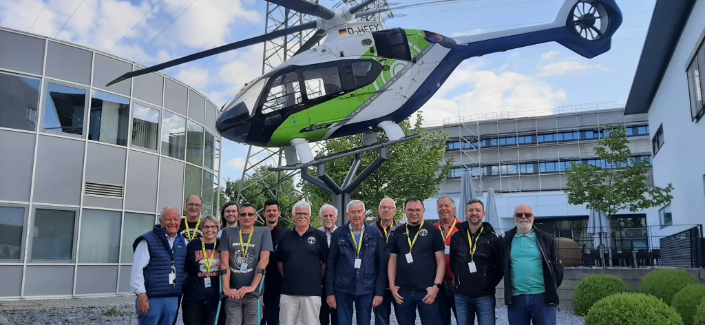 Gruppe von Männern vor einem grün-weiß-blauem Hubschraubermodell auf Metallgestell, Industriegebäude im Hintergrund