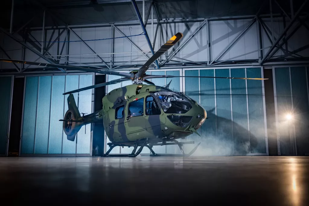 Grün-getarnter Militärhubschrauber vom Typ H145M in geöffnetem Hangar mit Nebel und Industriebeleuchtung