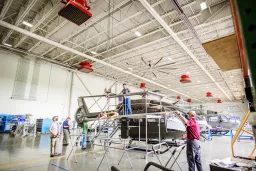 Blick in große Industriehalle mit Hubschrauber-Rohbau, mehrere Techniker an Arbeitsplattformen, rote Deckenlampen