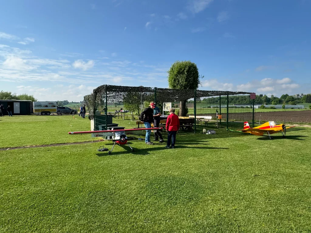 Grüne Rasenfläche mit zwei Modellflugzeugen, Flieger in roter und gelber Farbe, Zaun mit Netzabdeckung im Hintergrund