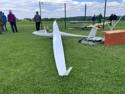 Weißes Segelflugmodell auf grüner Rasenfläche, im Hintergrund Modellflugplatz mit Zaun und Vereinsmitgliedern