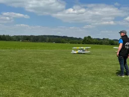 Weißes Doppeldecker-Modellflugzeug auf einer großen, gemähten Wiese mit Bäumen und blauem Himmel im Hintergrund