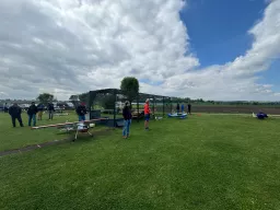 Grüne Rasenfläche mit Modellflugzeugen und mehreren Personen, Gitterzaun und Wolkenhimmel im Hintergrund