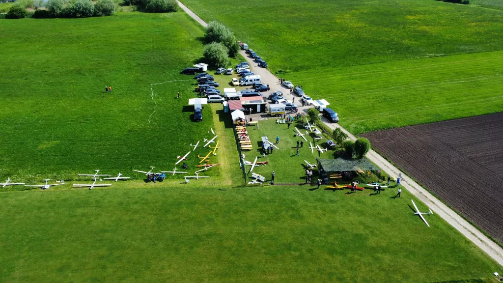 Luftaufnahme eines Modellflugplatzes mit weißen Segelflugzeugen, Autos und Zelten auf einer grünen Wiese neben einem Acker