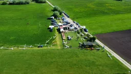 Luftaufnahme eines Modellflugplatzes mit weißen Segelflugzeugen, Autos und Zelten auf einer grünen Wiese neben einem Acker