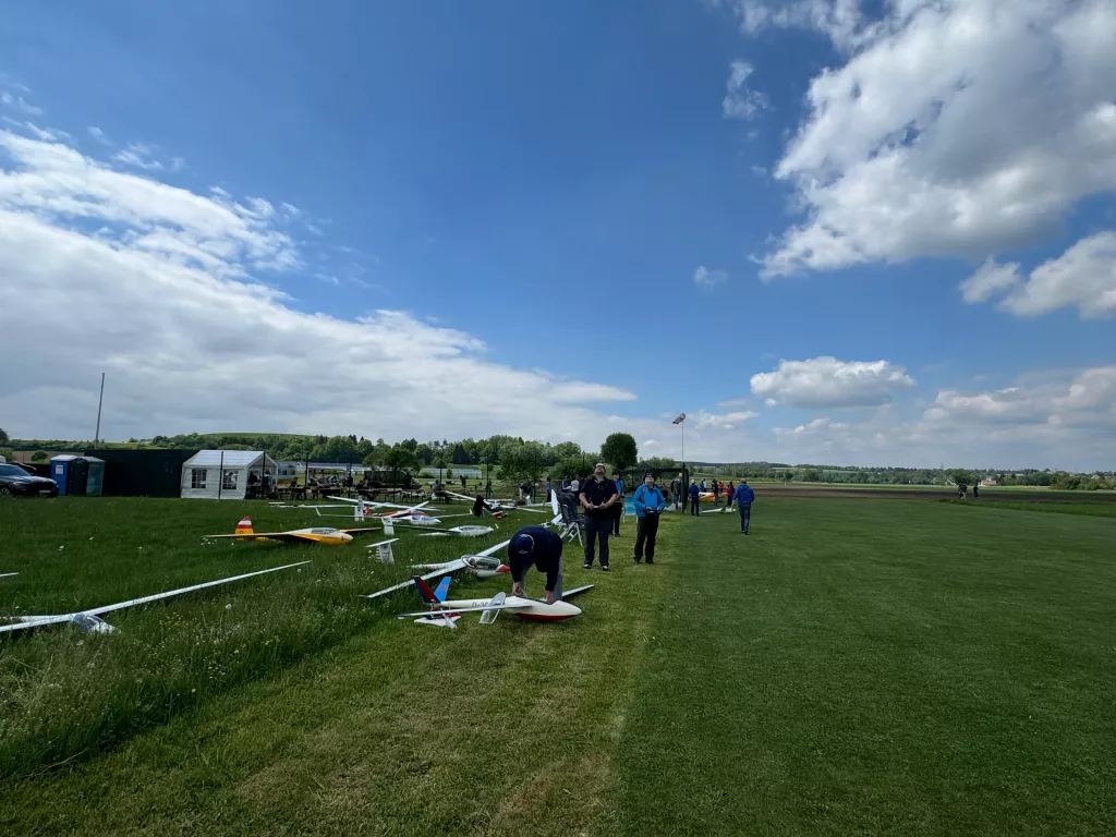 Grüne Wiese mit mehreren Modellflugzeugen, einige Personen im Hintergrund, blauer Himmel mit weißen Wolken