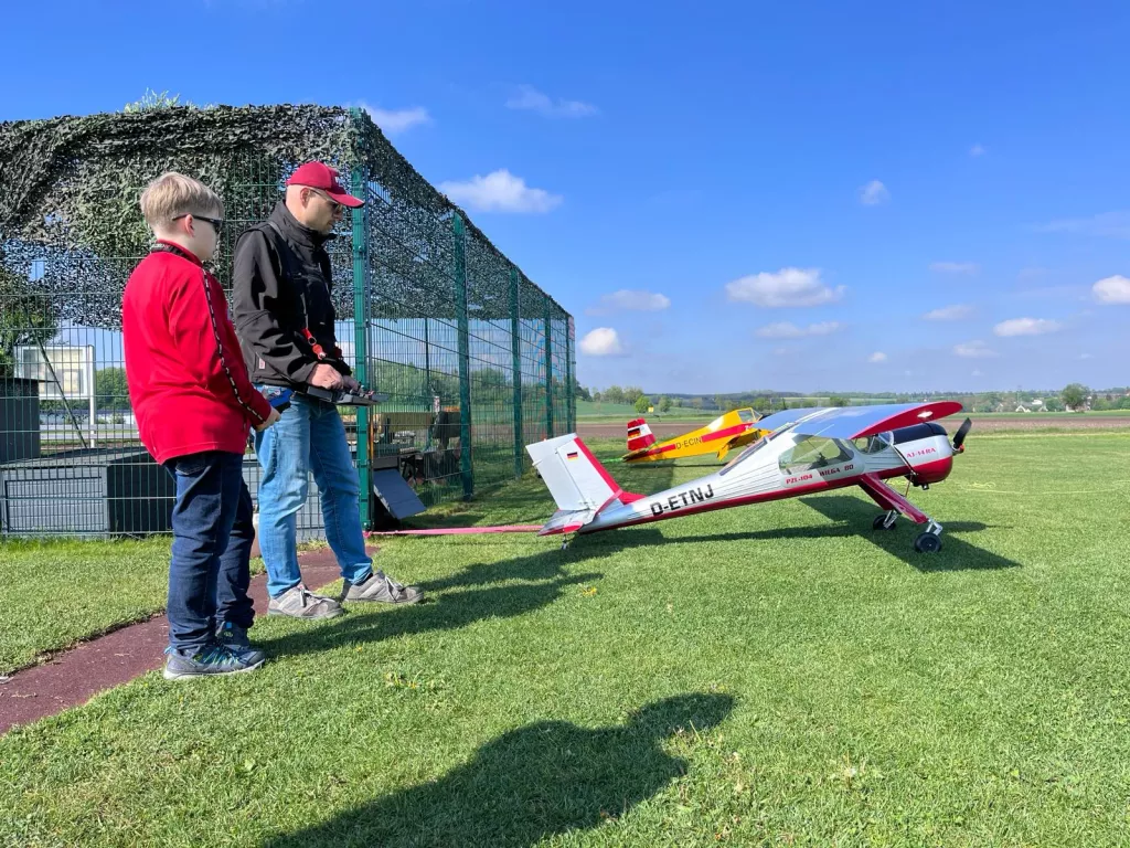 Zwei Personen an einem Modellflugplatz mit zwei roten Modellflugzeugen auf grünem Rasen, Zaun und blauem Himmel im Hintergrund
