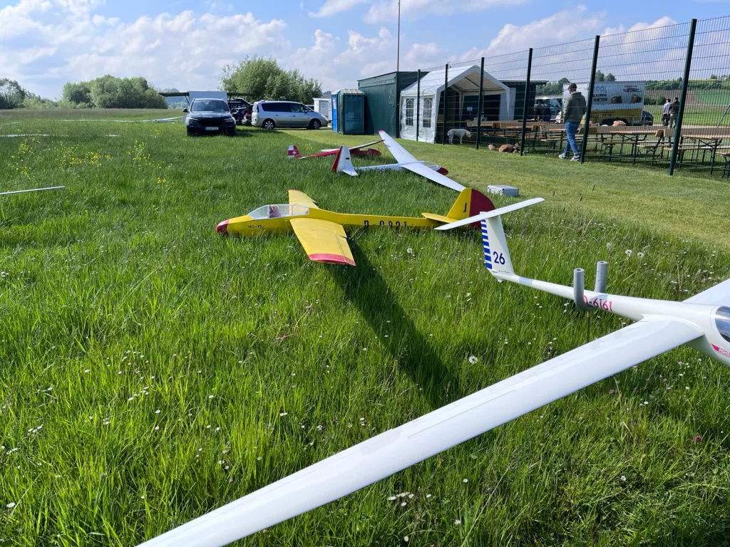 Drei Modellflugzeuge - gelb, weiß, rot - auf grüner Wiese mit Autos und Vereinsgebäuden im Hintergrund