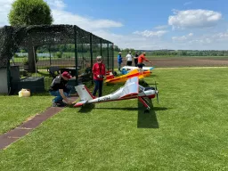 Zwei Modellflugzeuge auf einer Rasenfläche, Modellflieger in roter und blauer Jacke, sonniger Sommertag mit Feld im Hintergrund