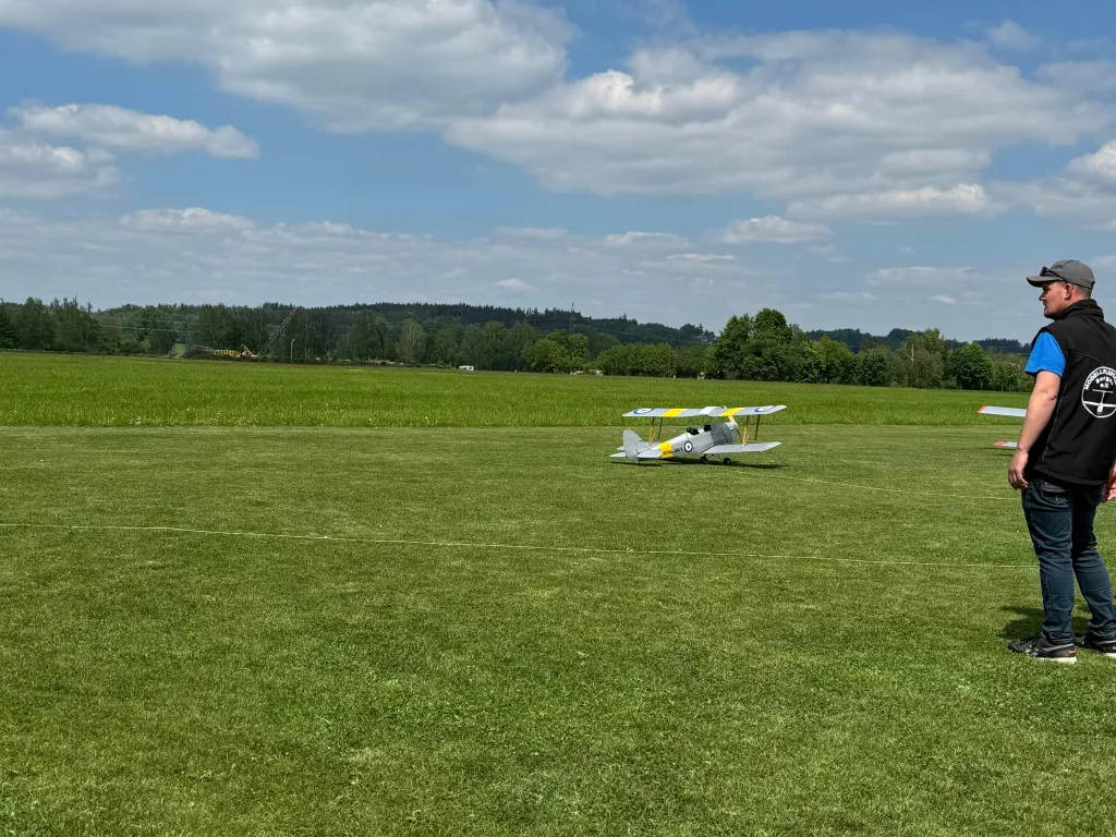 Weißes Doppeldecker-Modellflugzeug auf einer großen, gemähten Wiese mit Bäumen und blauem Himmel im Hintergrund