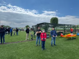 Mehrere Personen auf Grünfläche mit Modellflugzeugen, Netzunterstand, bewölkter Himmel, Vereinsgelände mit Modellfliegern