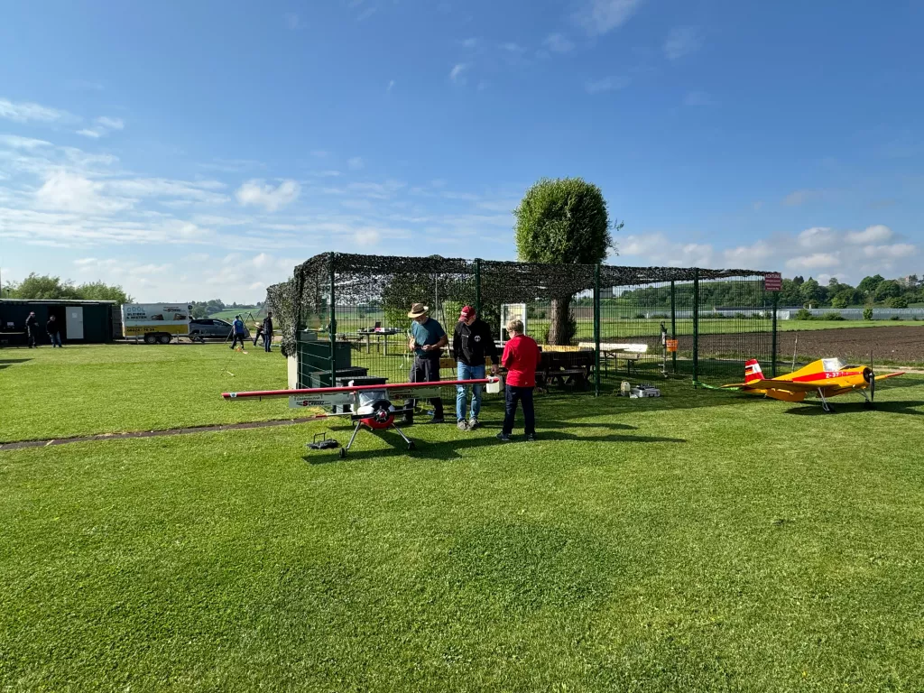 Grüne Wiese mit Modellflugzeugen, Zaun mit Tarnnetz, zwei Personen, blauer Himmel mit Wolken