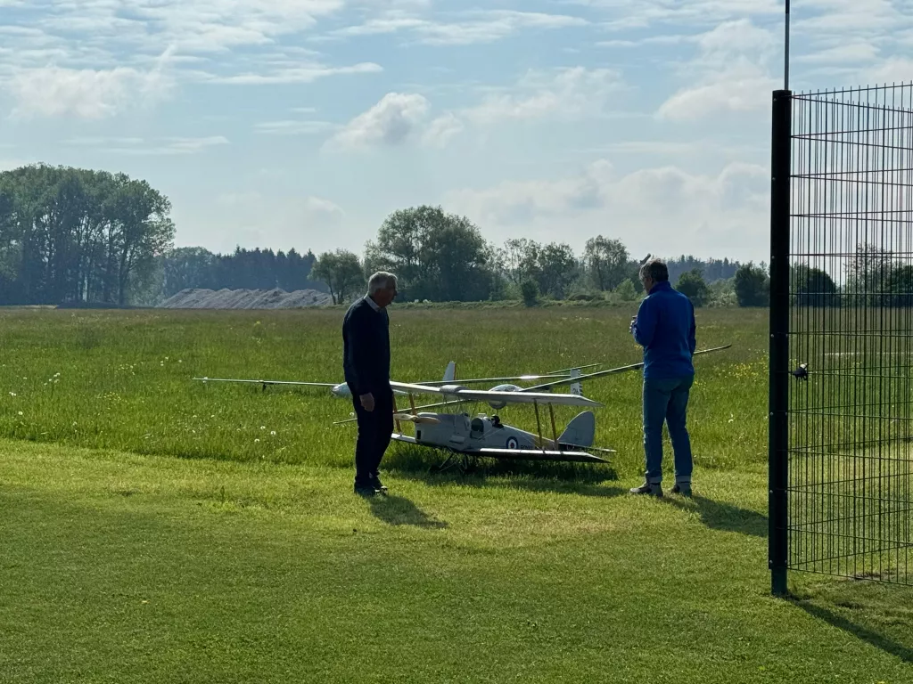 Zwei Personen stehen neben einem weißen Modellflugzeug auf einer grünen Wiese mit Bäumen im Hintergrund