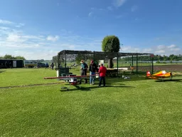 Grüne Wiese mit Modellflugzeugen, Zaun mit Tarnnetz, zwei Personen, blauer Himmel mit Wolken