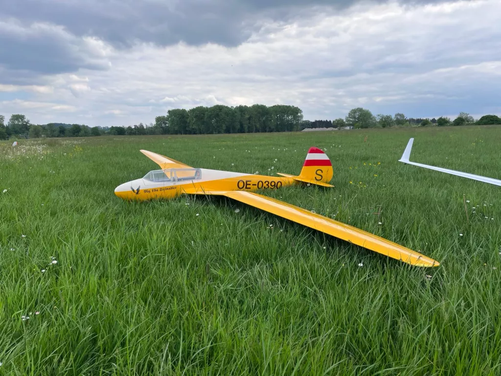 Gelbes Segelflugmodell mit Kennzeichen OE-0390 in saftig grüner Wiese, bewölkter Himmel im Hintergrund