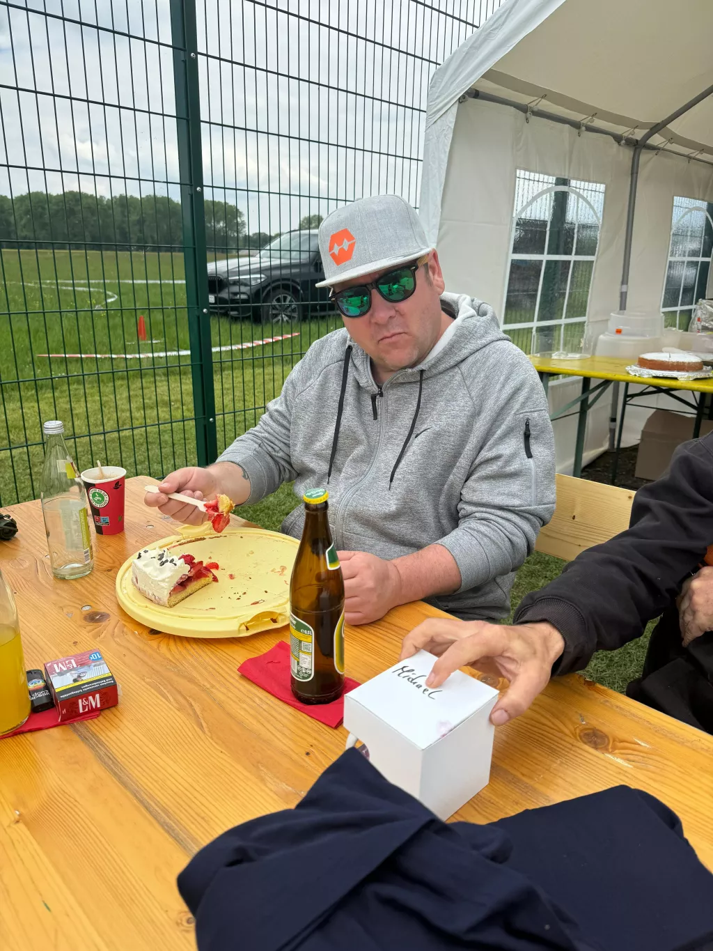 Mann mit Basecap und Sonnenbrille sitzt an Holztisch, isst Kuchen, Bier daneben, Zelt und Rasenplatz im Hintergrund