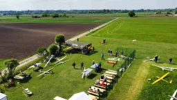 Luftaufnahme eines Modellflugplatzes mit mehreren weißen und gelben Segelflugmodellen auf grüner Wiese, umgeben von Feldern und Bäumen