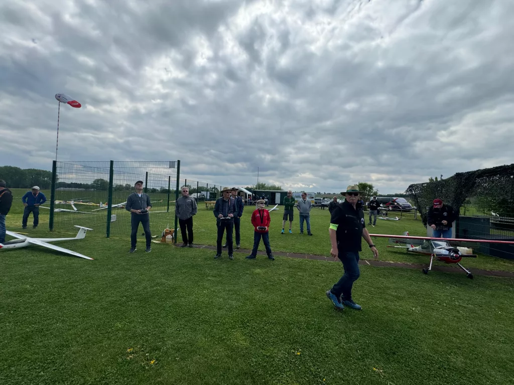 Modellflugplatz mit mehreren Personen, Segelflugmodellen und Zaun, grasbewachsene Fläche unter dramatischem Wolkenhimmel