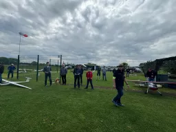 Modellflugplatz mit mehreren Personen, Segelflugmodellen und Zaun, grasbewachsene Fläche unter dramatischem Wolkenhimmel