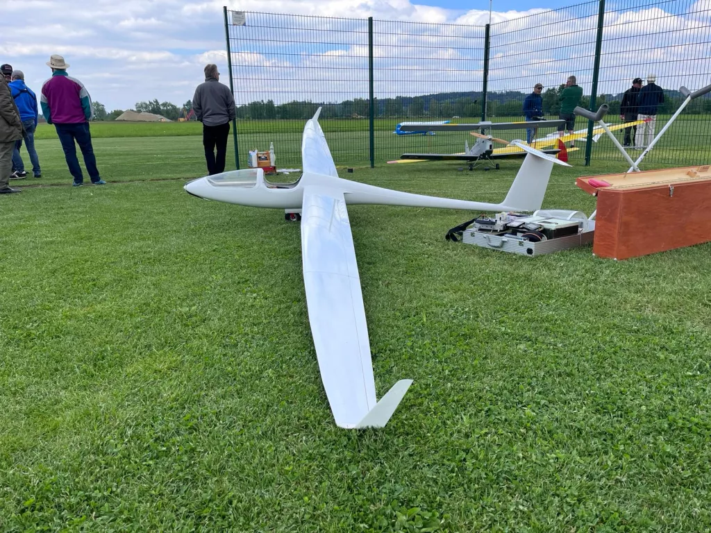 Weißes Segelflugmodell auf grüner Rasenfläche, im Hintergrund Modellflugplatz mit Zaun und Vereinsmitgliedern
