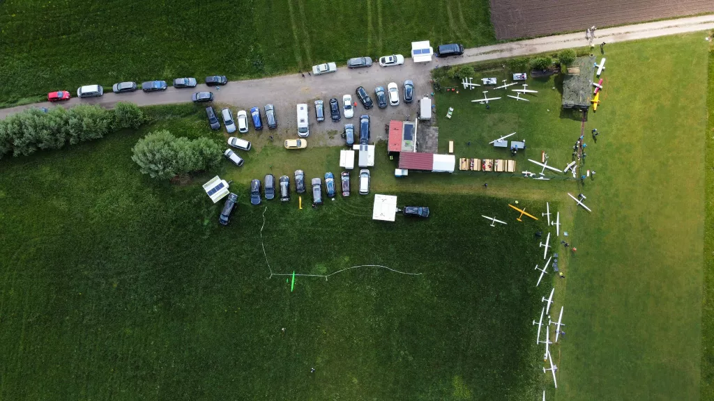 Luftaufnahme eines Modellflugplatzes mit mehreren weißen Segelflugmodellen und zahlreichen Autos auf Grünfläche