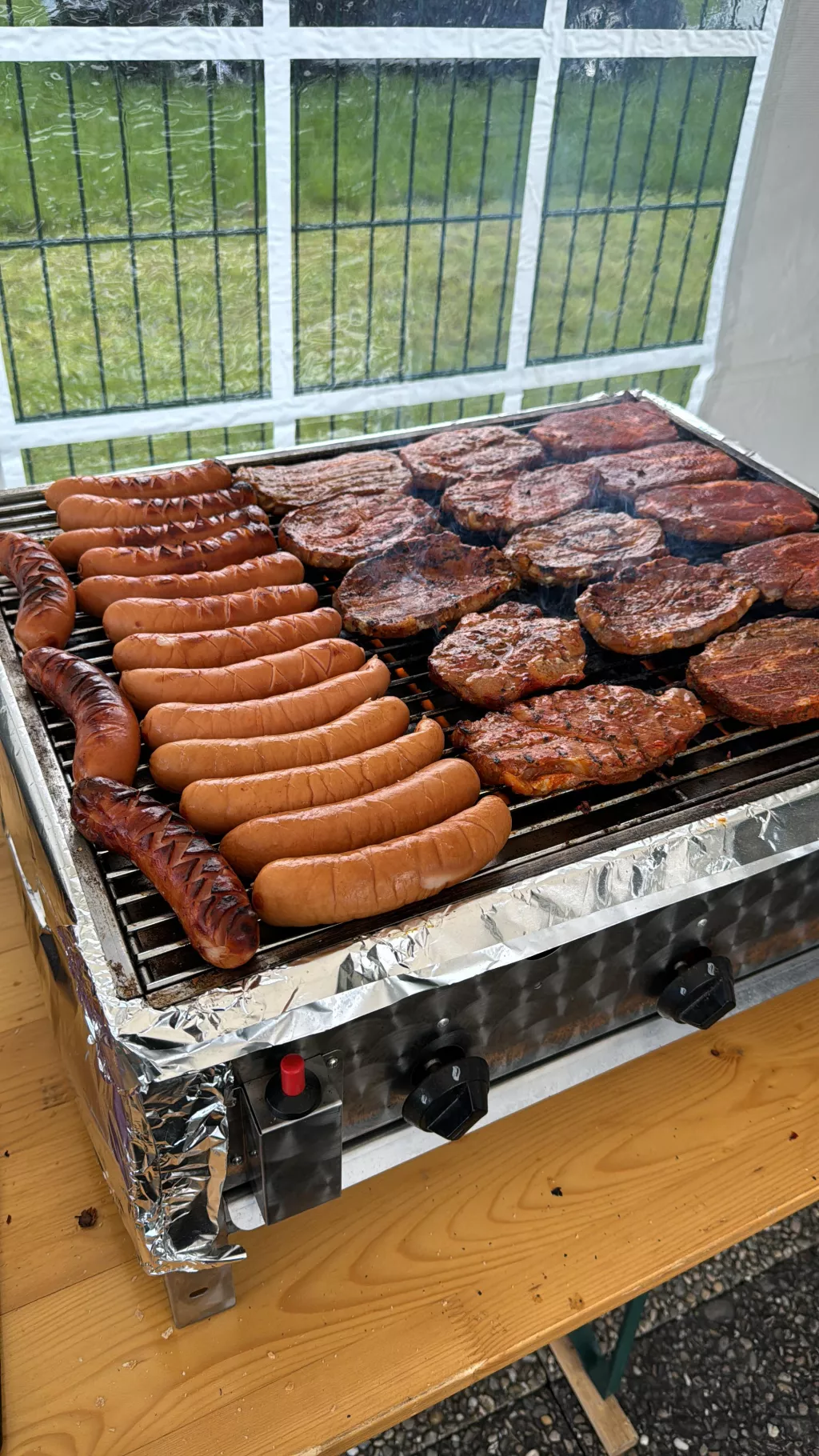 Elektrischer Tischgrill mit gebratenen Bratwürsten und Rindersteak auf Grillrost, Fenster im Hintergrund
