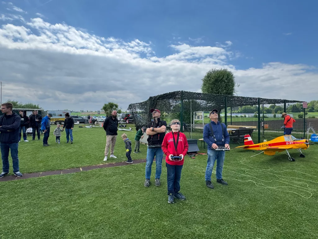 Mehrere Personen auf Grünfläche mit Modellflugzeugen, Netzunterstand, bewölkter Himmel, Vereinsgelände mit Modellfliegern