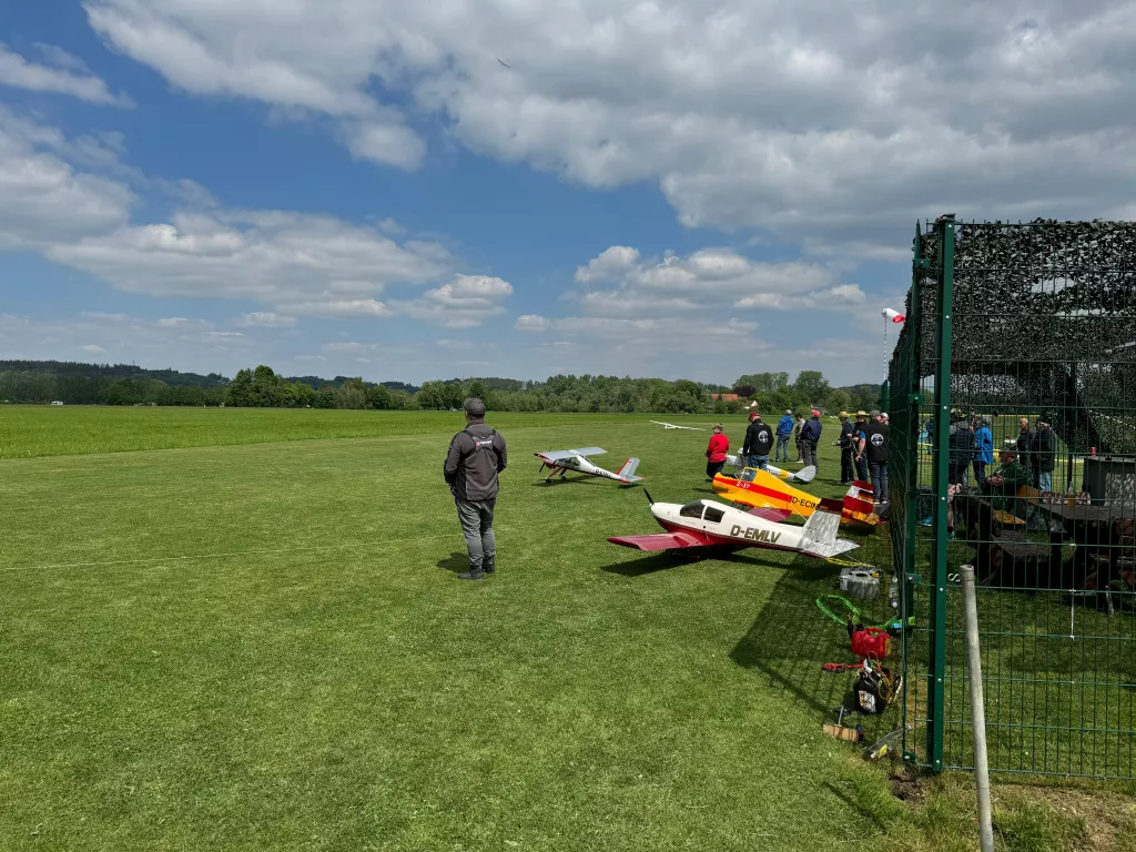 Mehrere Modellflugzeuge auf einer Wiese, Modellpiloten stehen hinter einem Zaun, bewölkter Himmel im Hintergrund