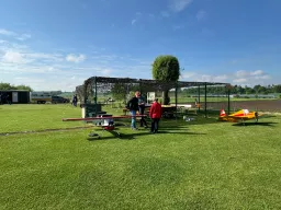 Grüne Rasenfläche mit zwei Modellflugzeugen, Flieger in roter und gelber Farbe, Zaun mit Netzabdeckung im Hintergrund