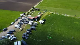 Luftaufnahme eines Modellflugplatzes mit mehreren Segelflugzeugen, Autos auf Parkplatz, grüner Wiese und Ackerfläche